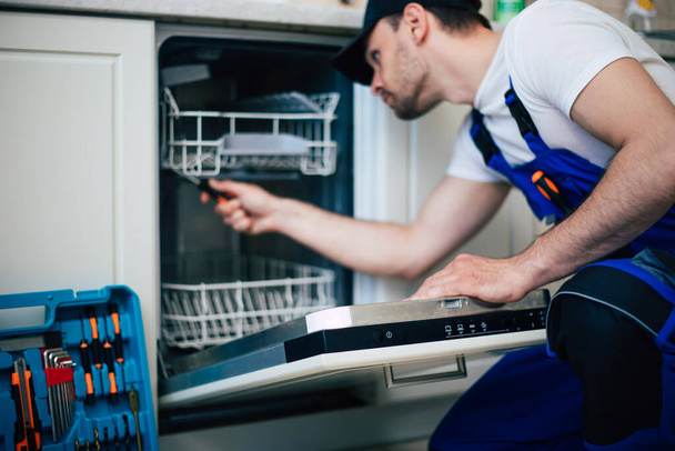 Young modern serviceman in worker suit during the repairing of the dishwasher on the domestic kitchen. - Photo, Image