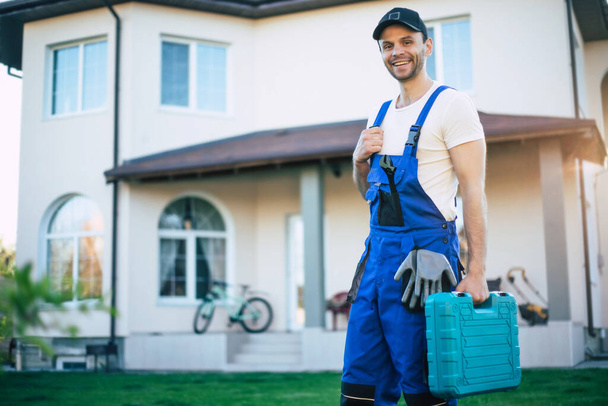 Handsome confident workman in special uniform with a modern toolbox is posing on a new big house background - 写真・画像