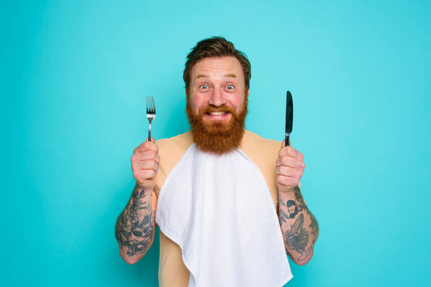 Щасливий чоловік з татуюваннями готовий їсти з столовими приборами в руці
 - Фото, зображення