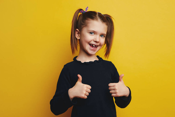 klein meisje met geopende mond tonen duimen omhoog geïsoleerd op gele muur - Foto, afbeelding