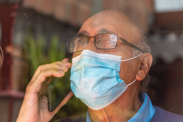 Ritratto di un vecchio anziano con occhiali e maschera medica che parla al telefono dietro un bicchiere con riflesso di edifici e cielo, mano sul vetro. Scena di pandemia di COVID o Coronavirus. - Foto, immagini