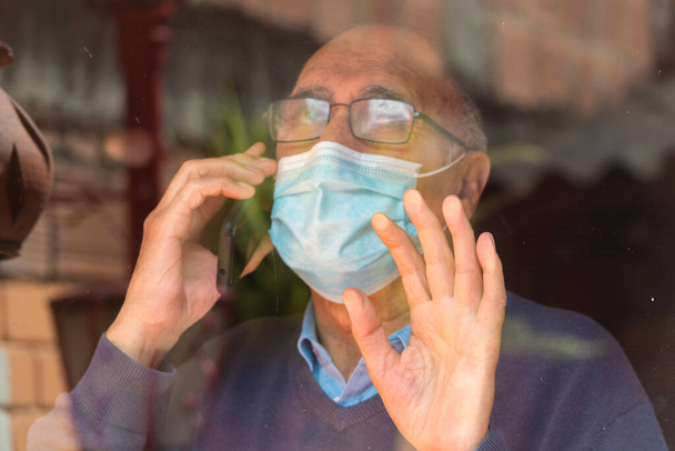 Muotokuva od vanha vanhempi mies lasit ja lääketieteellinen kasvonaamio puhuu puhelimessa lasin takana heijastus rakennusten ja taivaan, käsi lasi. COVID- tai Coronavirus-pandemian esiintyminen. - Valokuva, kuva