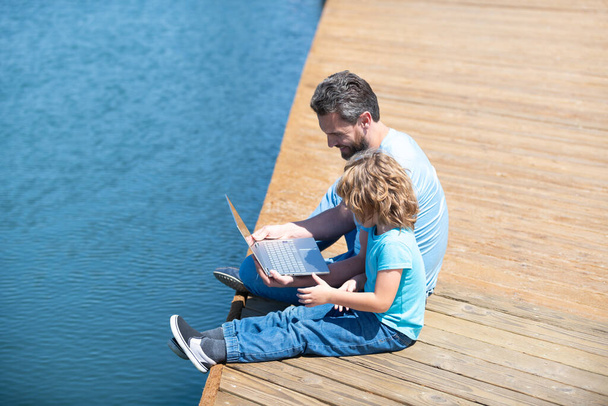 Πάρτε e-Learning σας στις καλοκαιρινές διακοπές. Πατέρας και γιος χρησιμοποιούν λάπτοπ. Συμμετοχή της οικογένειας στην ηλεκτρονική μάθηση - Φωτογραφία, εικόνα