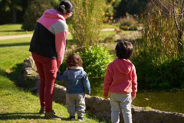 Matka z dwójką dzieci na plecach spacerująca po parku. Kobieta z dwójką dzieci w ogrodzie. Rodzinna sesja. Grupa osób - Zdjęcie, obraz