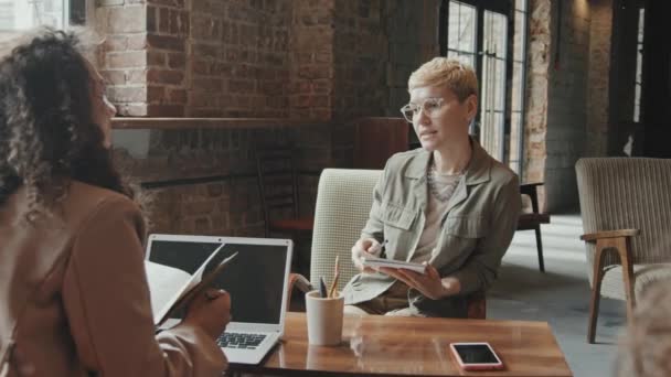 Rallentatore medio di giovane donna d'affari alla moda che discute il nuovo progetto con una collega seduta insieme alla scrivania in uno spazio di coworking in stile loft - Filmati, video