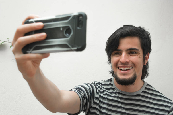 Portret młodego człowieka z brodą trzymającego telefon na dłoni uśmiechającego się i robiącego sobie selfie. Modelka w czarno-białej koszulce w paski. Szczęście i pewność siebie wyraz twarzy lub twarzy. - Zdjęcie, obraz