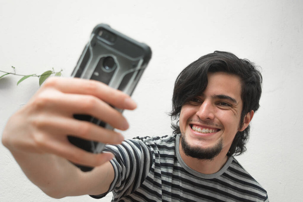 Portrét mladého muže s vousy, který drží telefon na ruce a usmívá se a bere si selfie. Model v černo-bílém pruhovaném tričku. Štěstí a sebevědomí obličeje nebo výrazů obličeje. - Fotografie, Obrázek