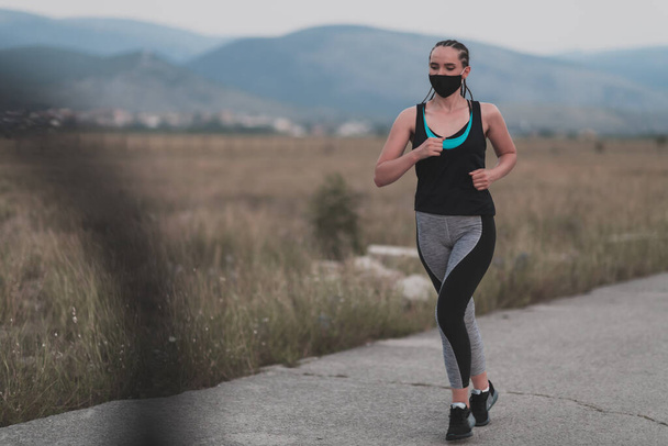 Determined fitness woman in short clothes wearing red protective face mask running outdoors in the city during coronavirus outbreak. Covid 19 and physical jogging activity, sport and fitness. - Photo, Image