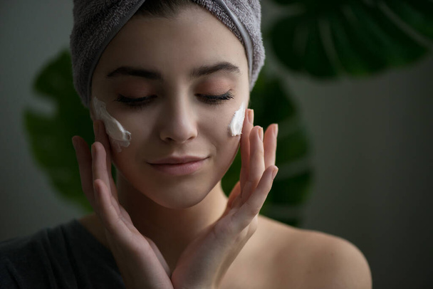 Young woman applying face cream on her face. Beauty model with perfect fresh skin and long eyelashes cares about her skin at home. Spa and Wellness, Skin Care Concept. Close up, selected focus. - Photo, image