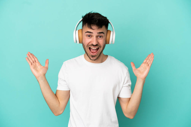 Young caucasian man isolated on blue background surprised and listening music - Photo, Image