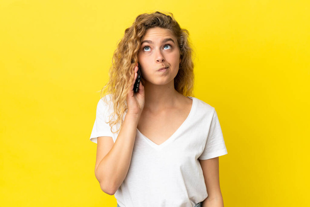 Young blonde woman using mobile phone isolated on yellow background and looking up - Photo, Image