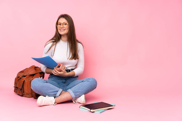 Teenager caucasian student girl sitting on the floor isolated on pink background with glasses and happy - Photo, Image