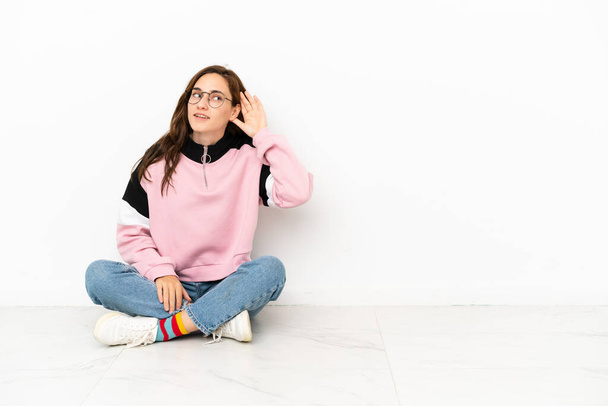 Young caucasian woman sitting on the floor isolated on white background listening to something by putting hand on the ear - Photo, Image