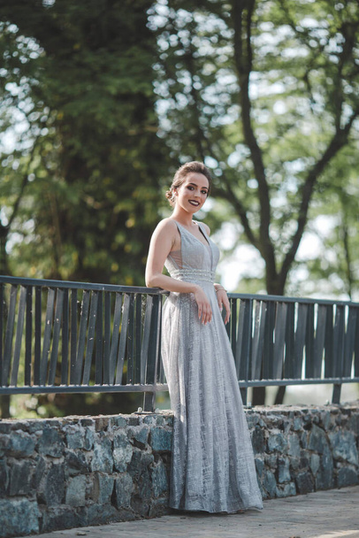 A portrait of a smiling Caucasian woman wearing a long gray dress and leaning on fences in a park - Foto, Imagem