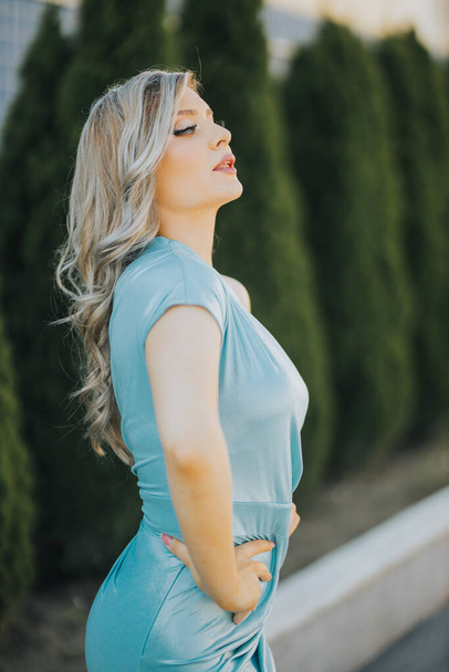 An attractive Caucasian young female in a baby blue dress posing outside next to spruce trees - Foto, afbeelding