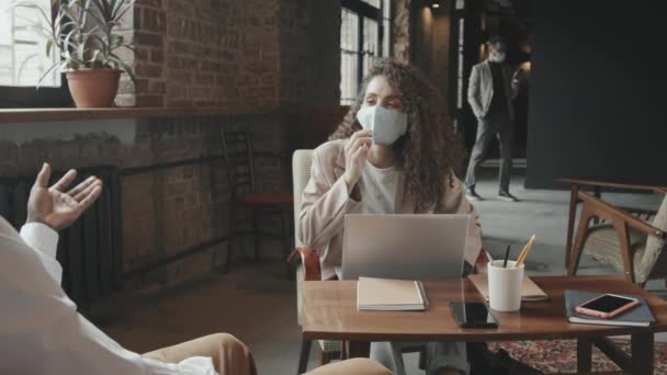 Medium traagheid van aantrekkelijke jonge zakenvrouw in gezicht masker praten met haar Afro-Amerikaanse zakenpartner zitten aan tafel in moderne loft coworking ruimte - Video