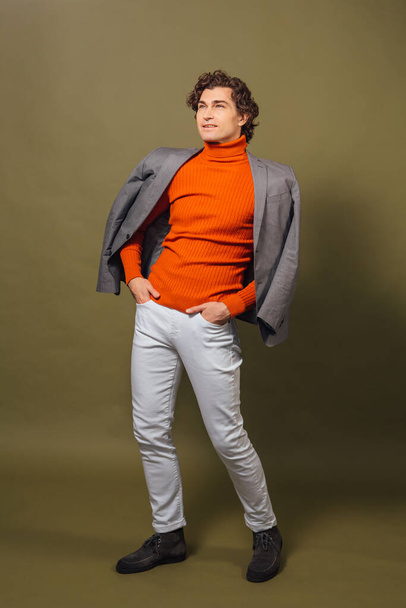 Full length portrait of a tall handsome man dressed in orange turtleneck, grey jacket and white jeans posing on the green background - Foto, afbeelding