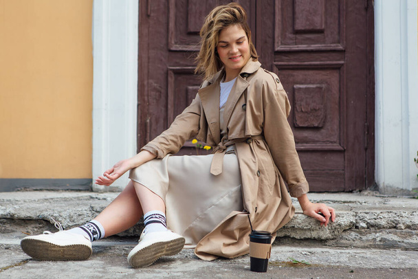 Young smiling millennial woman with wild hair dressed in an autumn coat sitting near the door of an old building with a cup of coffee to go. - Zdjęcie, obraz