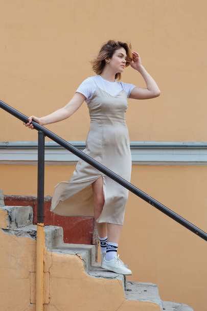Young smiling millennial woman with wild hair dressed in a beige dress posing on the stairs of an old building. - Foto, Imagem