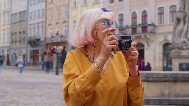 Mujer mayor turista tomando fotos con cámara fotográfica en el centro de la ciudad de verano, la vida después de la jubilación - Imágenes, Vídeo