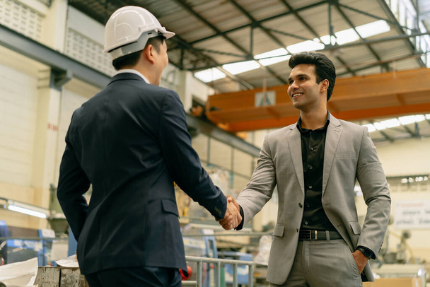30s young Asian businessman in formal suit and hard hat showing foreign business partner in factory background. Business partnership and team work success concept - Foto, Imagen