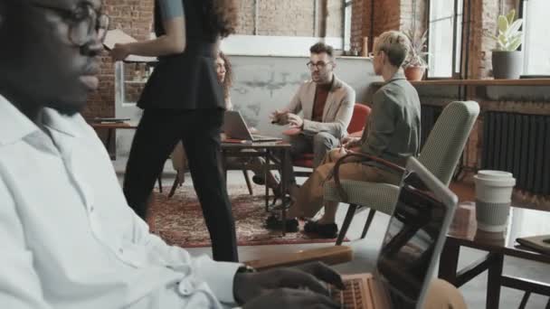 Zoom-in slowmo van diverse zakenmensen die werken in moderne loft-stijl coworking ruimte Afro-Amerikaanse man typen op laptop, terwijl drie zakelijke partners bespreken project aan de volgende tafel - Video
