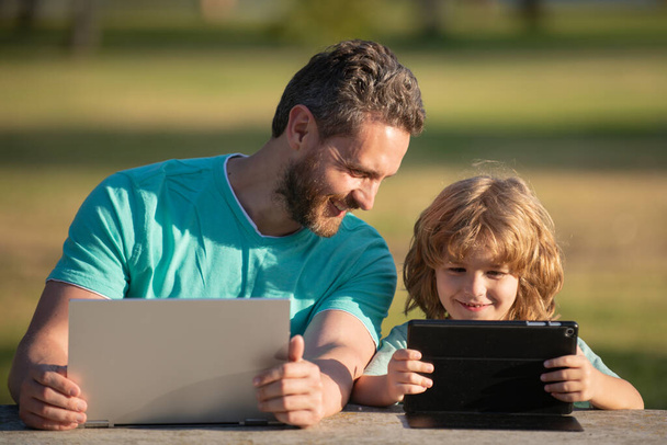 Father teaching son to use laptop, dad and school boy child looking computer screen and tablet, playing game, watching video, sitting on grass. Outdoor family weekend. - Photo, Image