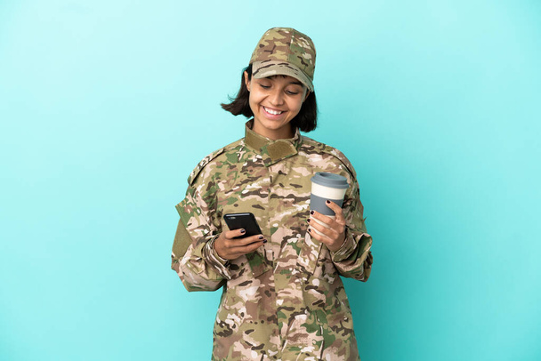 青い背景に隔離された軍事混合人種の女性がコーヒーを持ち去り、携帯電話を持って - 写真・画像
