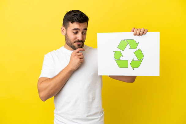 Young handsome caucasian man isolated on yellow background holding a placard with recycle icon - Foto, imagen