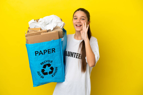 Mała dziewczynka trzyma torbę z makulaturą pełną papieru do recyklingu na odizolowanym żółtym tle krzycząc z otwartymi ustami - Zdjęcie, obraz