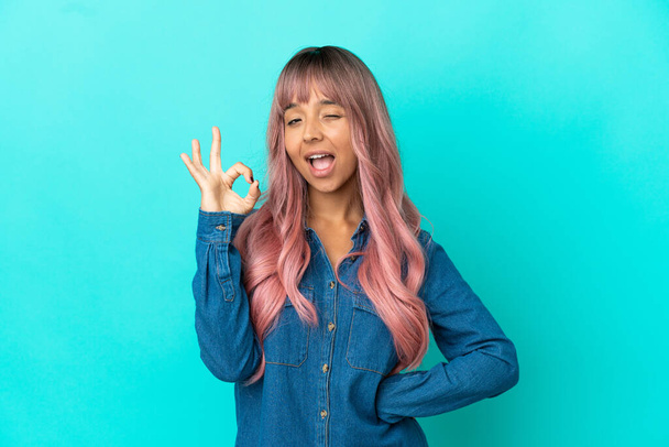 Jovem mulher de raça mista com cabelo rosa isolado no fundo azul mostrando sinal ok com os dedos - Foto, Imagem