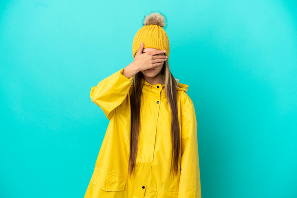 Menina vestindo um casaco à prova de chuva sobre fundo azul isolado cobrindo os olhos por mãos. Não quero ver nada. - Foto, Imagem