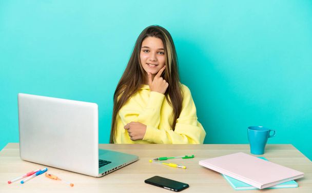 Kislány egy asztalnál egy laptop felett elszigetelt kék háttér boldog és mosolygós - Fotó, kép