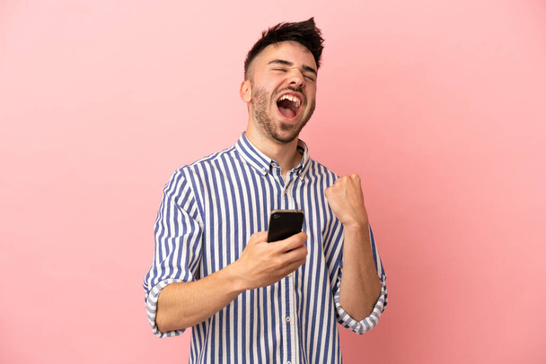 Mladý kavkazský muž izolované na růžovém pozadí s telefonem ve vítězné pozici - Fotografie, Obrázek