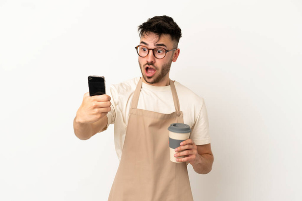 Εστιατόριο σερβιτόρος καυκάσιος άνθρωπος απομονώνονται σε λευκό φόντο κρατώντας καφέ για να πάρει μακριά και ένα κινητό - Φωτογραφία, εικόνα