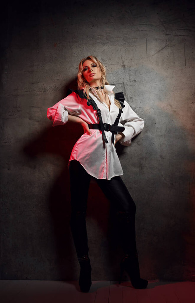 Insolent blonde woman mistress in leather pants, white shirt with leather belts and cap stands at concrete wall - Photo, image
