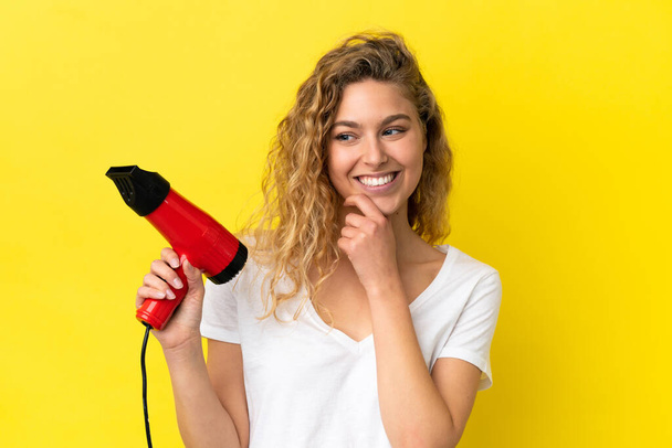 Sarı arka planda izole edilmiş saç kurutma makinesini tutan genç sarışın kadın yana bakıyor ve gülümsüyor. - Fotoğraf, Görsel