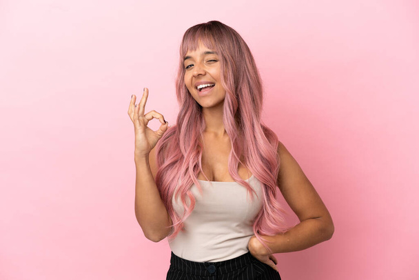 Jovem mulher de raça mista com cabelo rosa isolado no fundo rosa mostrando sinal ok com os dedos - Foto, Imagem