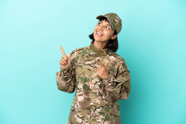 Военная женщина смешанной расы изолированы на синем фоне указывая указательным пальцем отличная идея - Фото, изображение