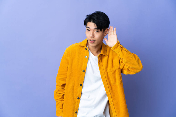Young Chinese man isolated on purple background listening to something by putting hand on the ear - Фото, изображение