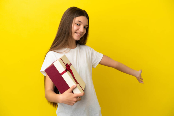Bambina che tiene un regalo su sfondo giallo isolato tendendo le mani di lato per invitare a venire - Foto, immagini