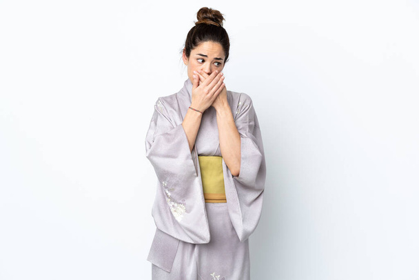 Frau trägt Kimono über isoliertem Hintergrund, der den Mund bedeckt und zur Seite schaut - Foto, Bild
