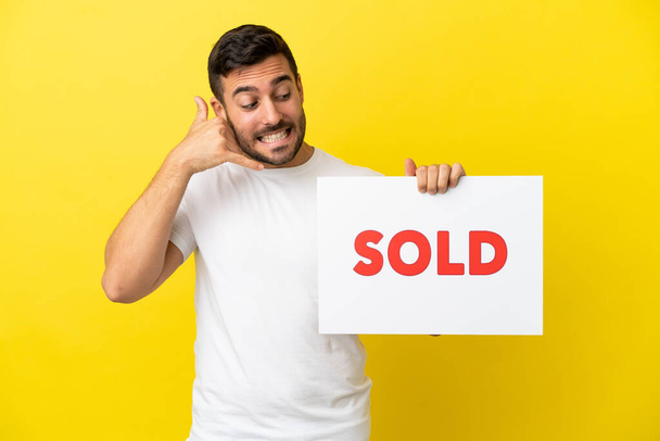Young handsome caucasian man isolated on yellow background holding a placard with text SOLD and doing coming gesture - Fotó, kép