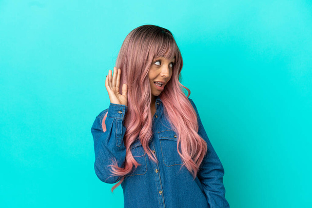 Nuori sekoitettu rotu nainen vaaleanpunaiset hiukset eristetty sinisellä taustalla kuuntelemalla jotain laittamalla käsi korvaan - Valokuva, kuva