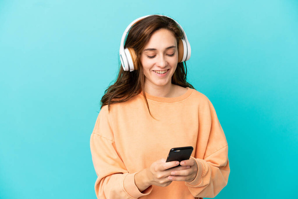 Jovem caucasiana isolada em fundo azul ouvindo música e olhando para celular - Foto, Imagem