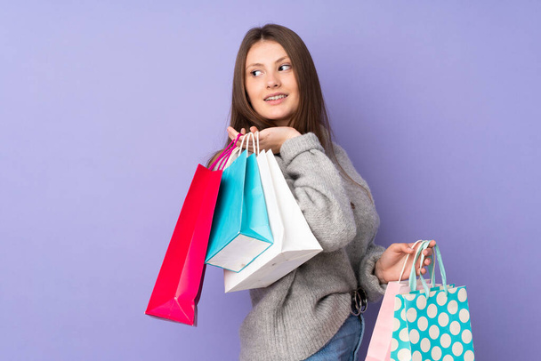 Adolescente caucásico chica aislado en púrpura fondo sosteniendo bolsas de compras y sonriendo - Foto, imagen