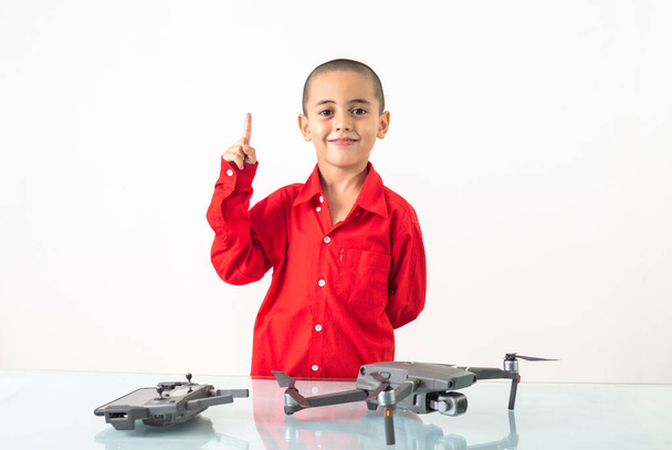 Mignon garçon avec coiffure skinhead dans une chemise rouge à manches longues assemble un drone sur une table en verre clair avec une toile de fond blanche - Photo, image