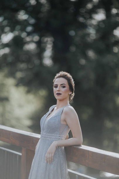 A portrait of a brunette Caucasian woman wearing a long gray dress and leaning on fences in a park - Foto, Imagen
