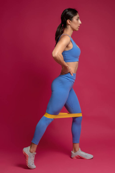 Young woman performing leg training with resistance band on maroon background - Foto, imagen
