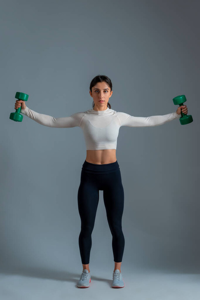 Chica realizando conjunto de ejercicios de hombro con mancuernas sobre fondo gris - Foto, Imagen